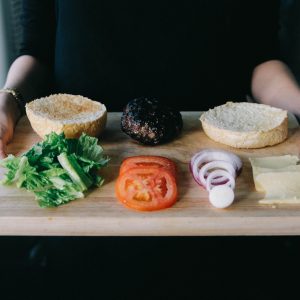 woman holding up wooden tray with lettuce, tomato, onions rings and cooked patty for burger